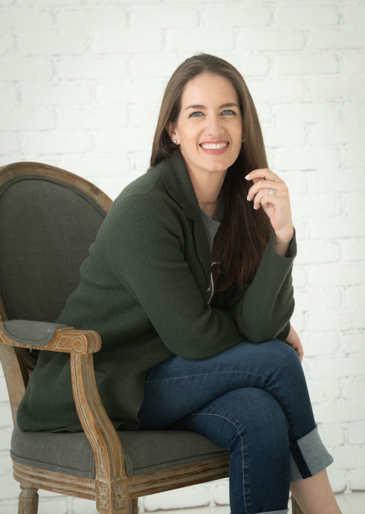 smiling woman professional natural light studio photo of woman sitting in chair wearing green sweater legs crossed with hand up to face and smiling off camera dallas texas studio rental